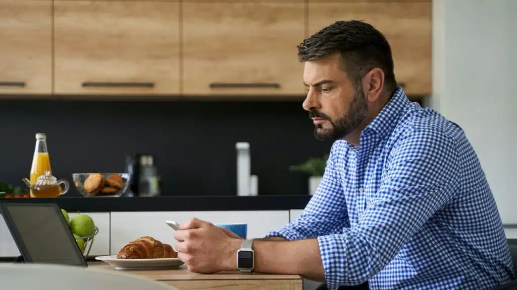 A focused man with a beard sits at a kitchen table, looking at his smartphone with a croissant and tablet nearby.