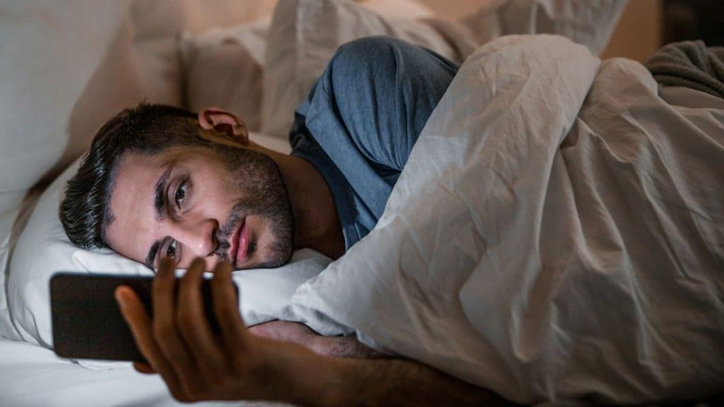 A bearded man lies in bed, under a white comforter, looking intently at a dark-screened smartphone.