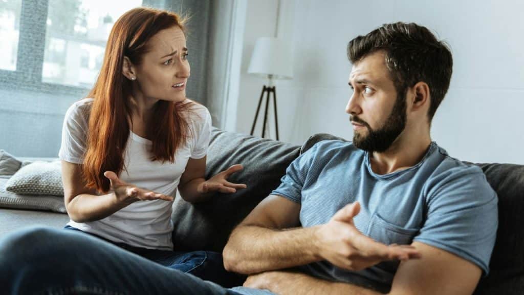 A frustrated red-haired woman sits on a couch, gesturing at a bearded man who is looking at her with a confused expression.