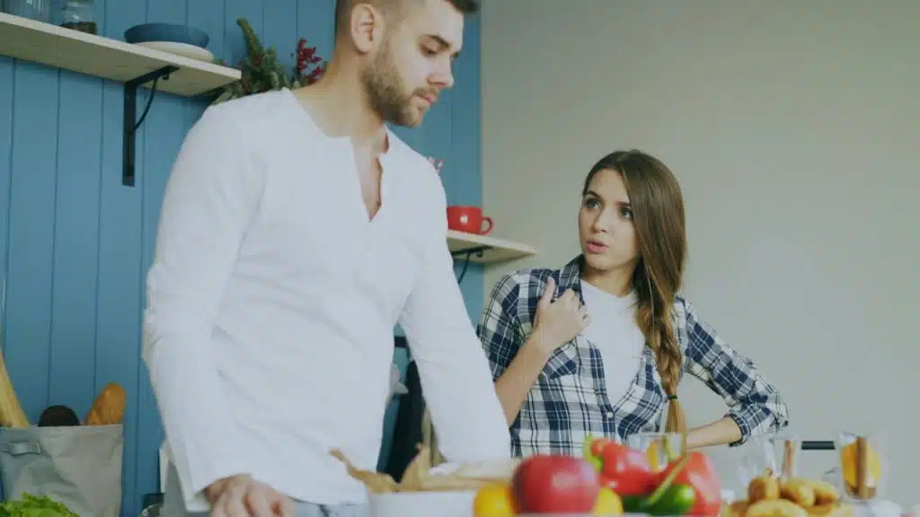 A man and a woman are standing in a kitchen with a frustrated expression on the woman's face.
