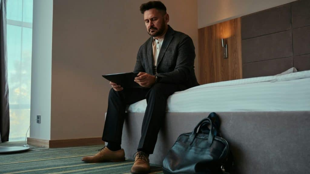 A serious-looking man sits on a bed, holding a tablet.