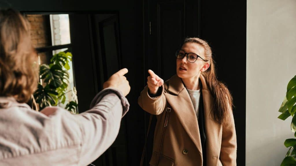 A woman in a tan coat and glasses angrily points at a person with their back to the camera.