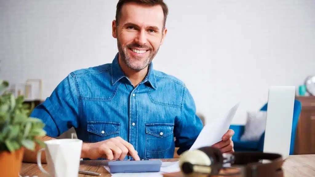 Smiling man in a denim shirt using a calculator and holding a paper.