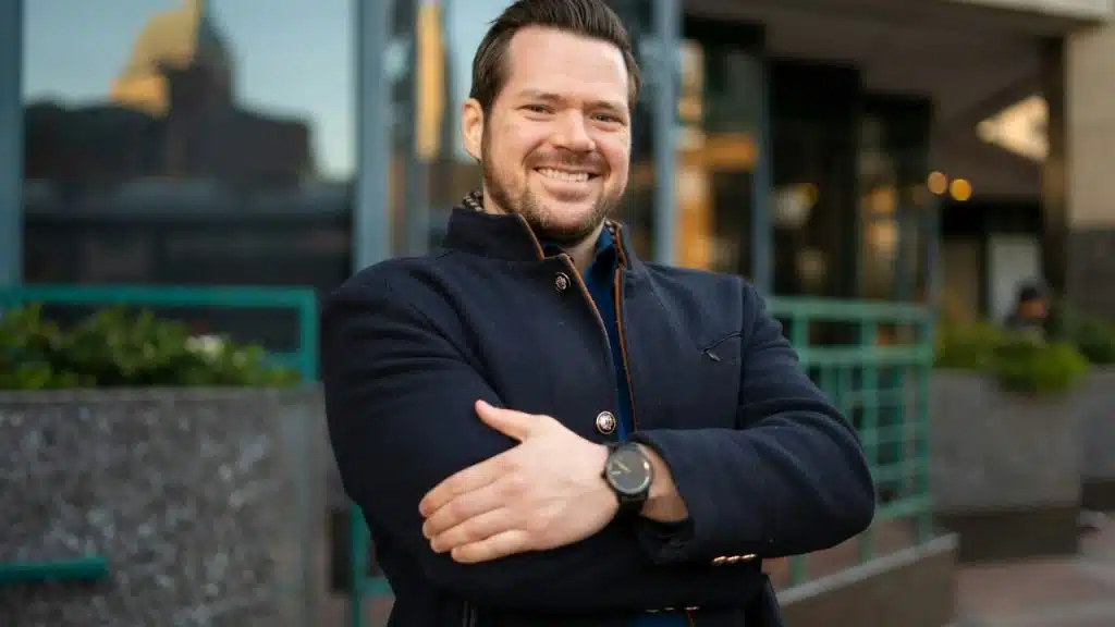 Smiling man in a dark coat with arms crossed standing outside an office building.