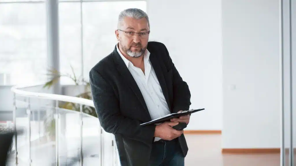 Older man in glasses, blazer, and white shirt holding a clipboard indoors.