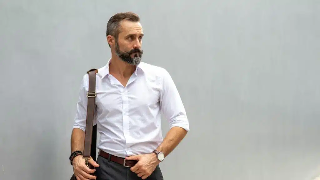 A bearded man in a white shirt with a bag on his shoulder.