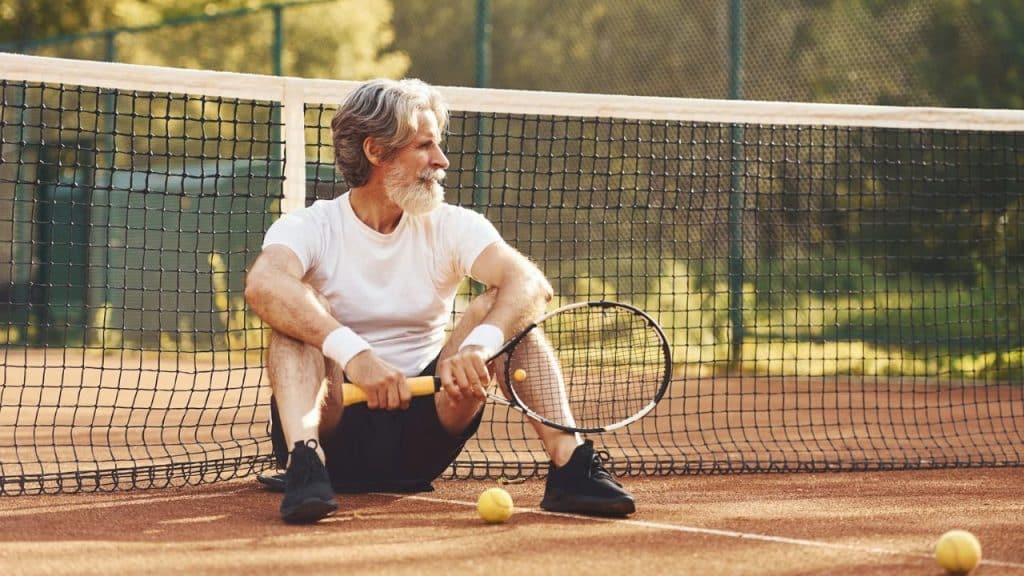 Senior man with white beard sits on a clay tennis court, holding a racket.