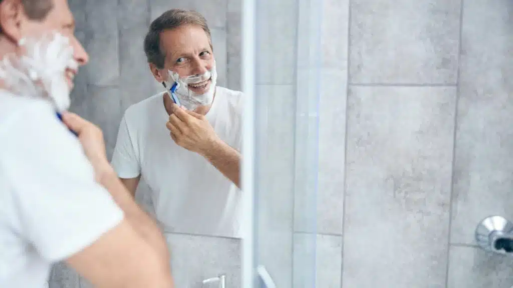 Mature man in white shirt shaving his face with cream in a bathroom mirror.