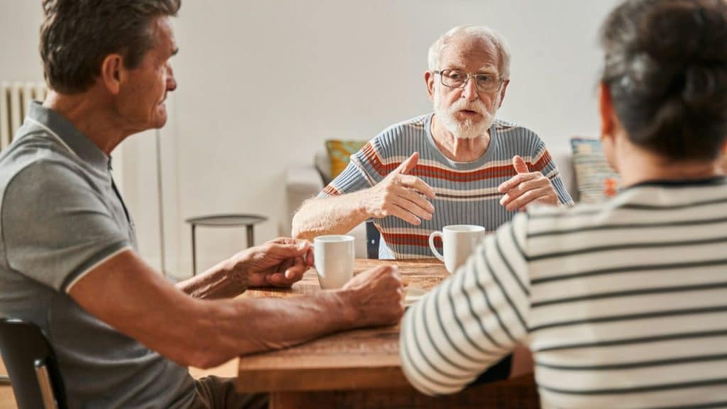 Three seniors sit at a wooden table, one man speaks, gesturing with his hands.