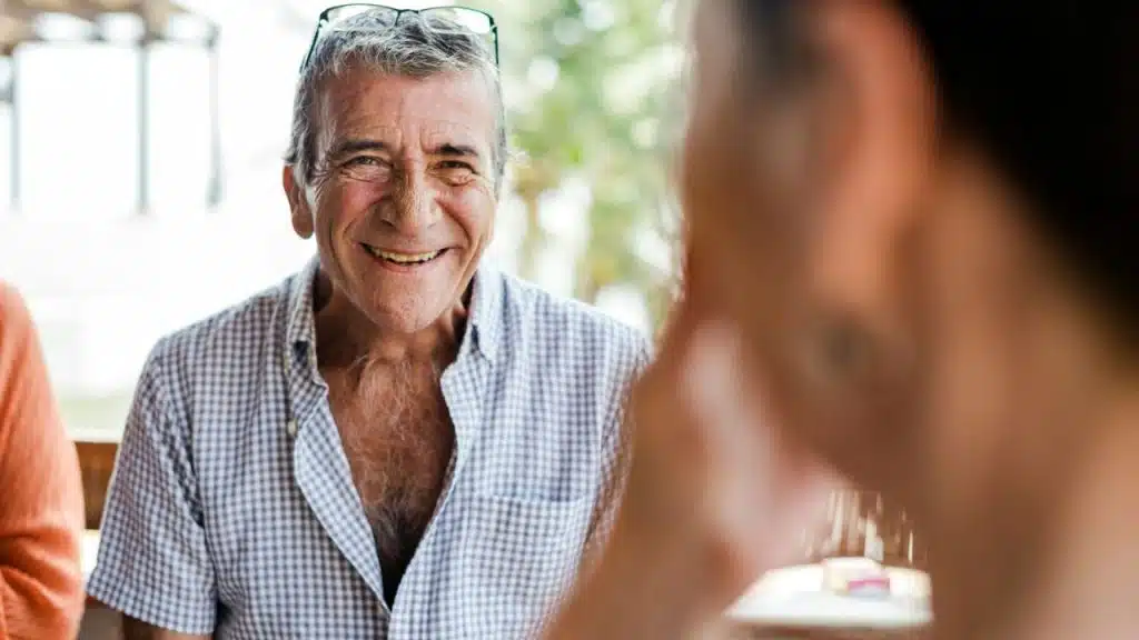 Smiling older man in a checkered shirt, with glasses on his head, facing a person.