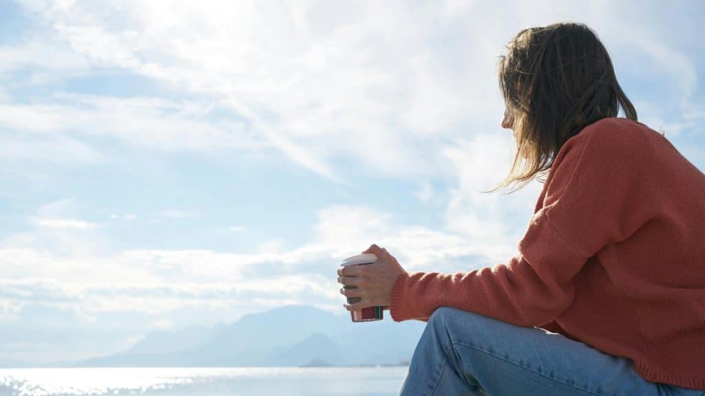A woman sitting by the water holding a cup and looking at the view.