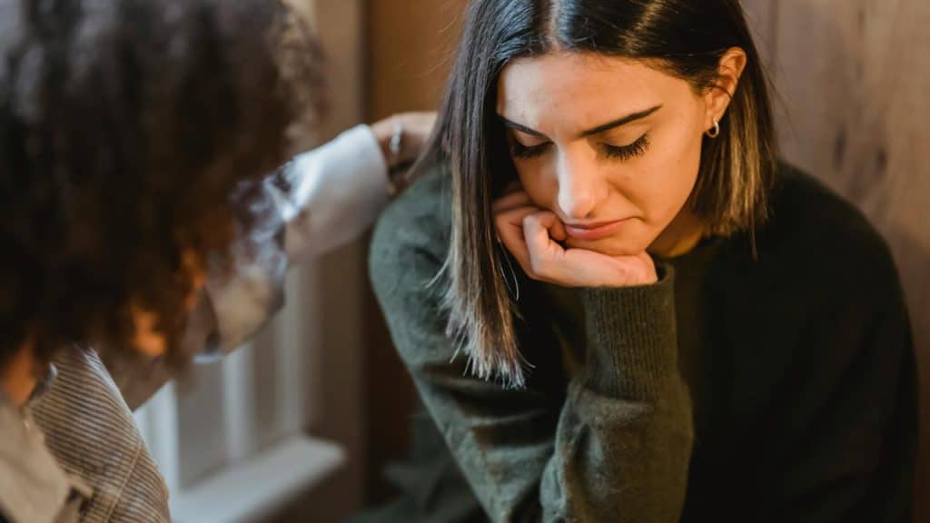 A woman looking down sadly while a friend comforts her with a hand on her shoulder.