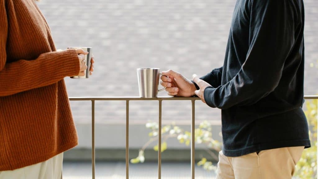 A couple standing on a balcony holding coffee mugs and talking.