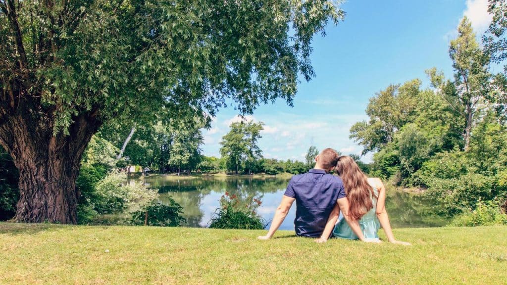 A couple sitting on the grass by a lake, leaning on each other.