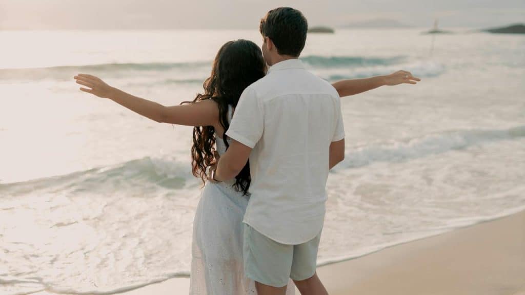 A couple standing on the beach with their arms outstretched, watching the waves.