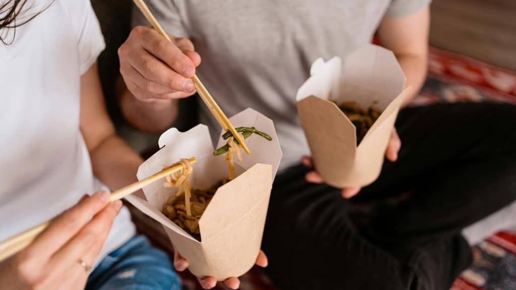 A couple eating takeout noodles with chopsticks while sitting together.