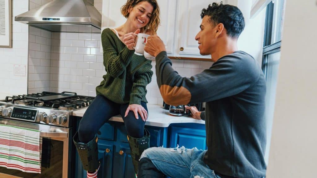 A couple smiling and clinking coffee mugs together in a kitchen.