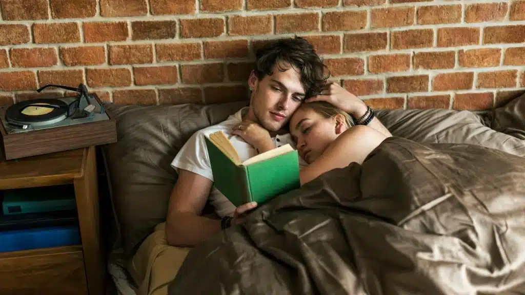 A couple lying in bed together while one reads a book.