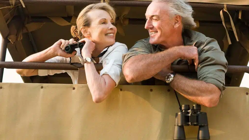 A mature couple smiling and chatting while holding binoculars on a safari vehicle.