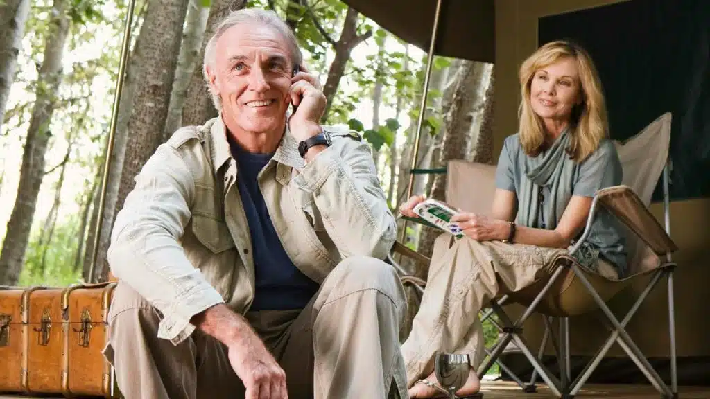 A mature man talking over the phone while a woman sitting behind looks at him.