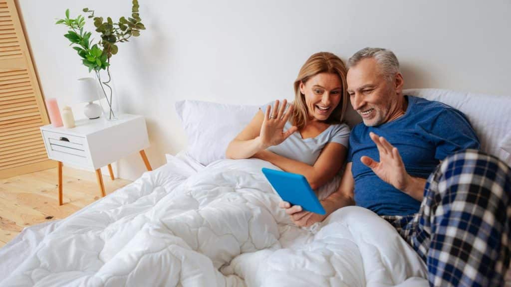 A mature couple waving their hands to a person they’re talking to on a tablet.