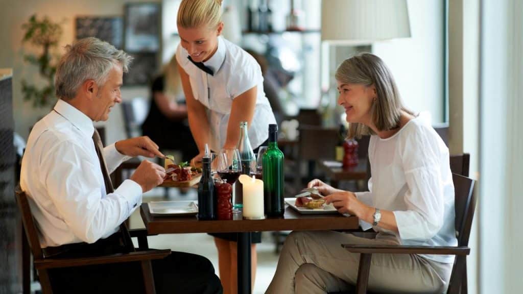 A female waiter serving food and drinks to a mature couple.