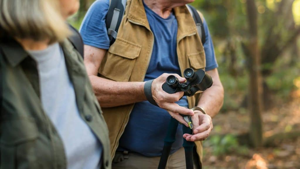 A person holding a pair of binoculars out on the woods with another person beside them.