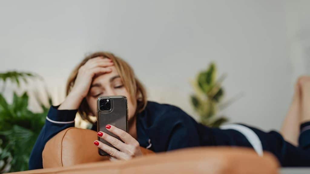 A woman lying on a couch looking at her phone with a hand on her forehead.