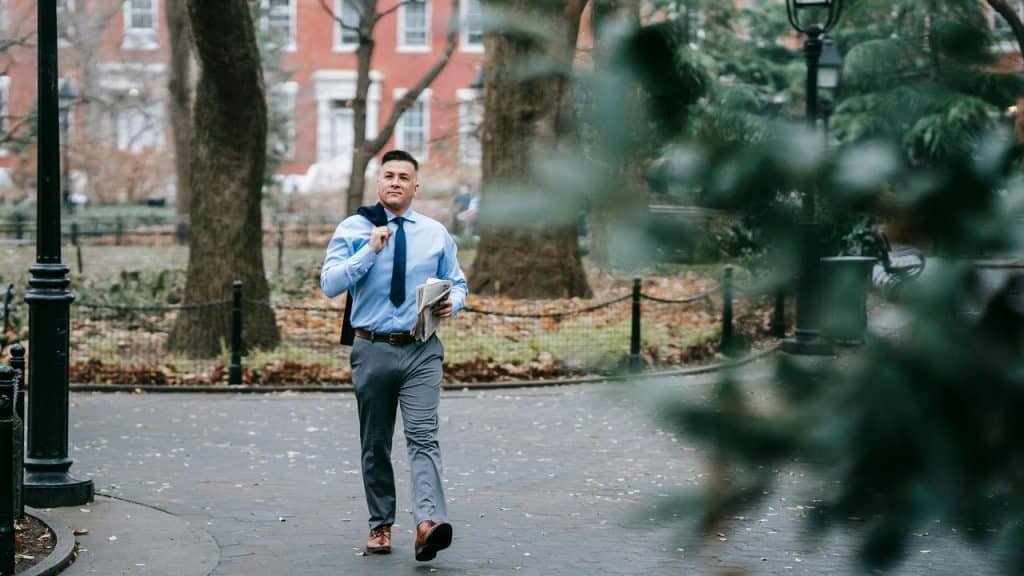 A man walking through a park holding a newspaper and jacket.