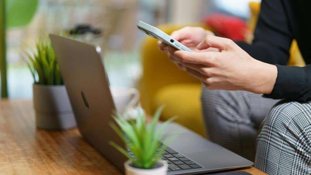 A person sitting at a table using a smartphone beside an open laptop.