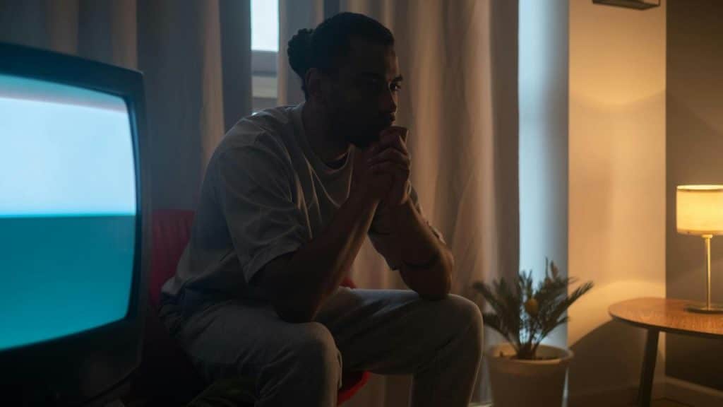 A man sitting in a dimly lit room with his hands clasped, looking thoughtful.