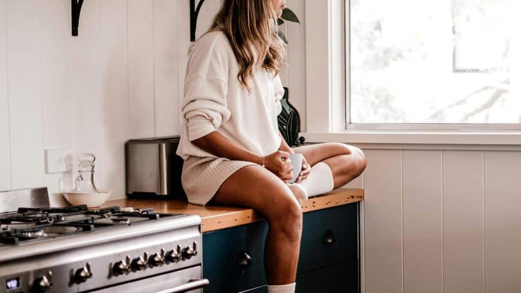 A woman sitting on a kitchen counter holding a cup and looking out the window.