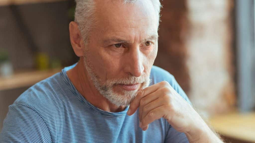 Close-up of an older man with gray hair and a beard, looking downward with a pensive expression.