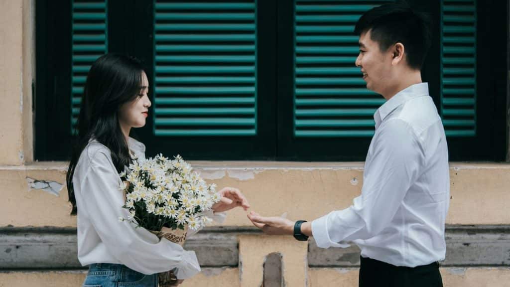 A smiling man in a white shirt reaches out to a woman who holds a large bouquet of white flowers.
