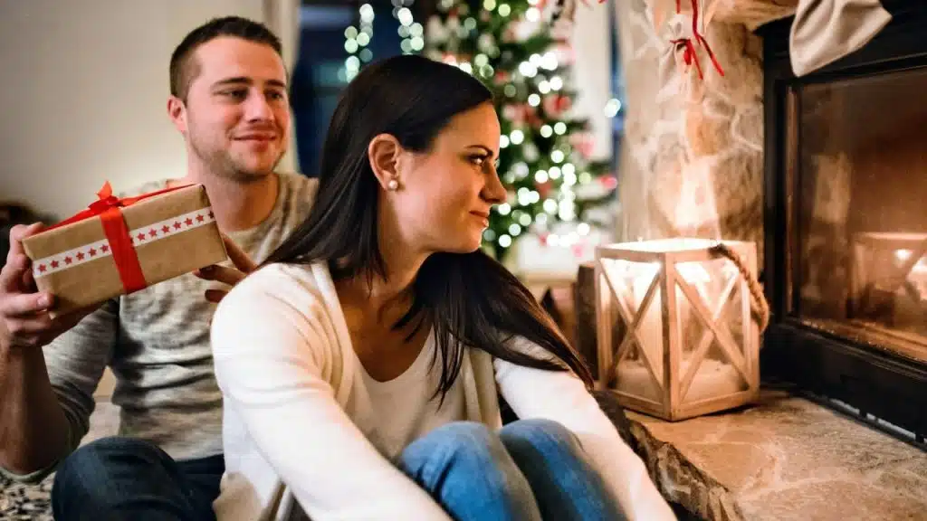 A man smiles while holding a wrapped gift behind a woman who looks towards a fireplace.