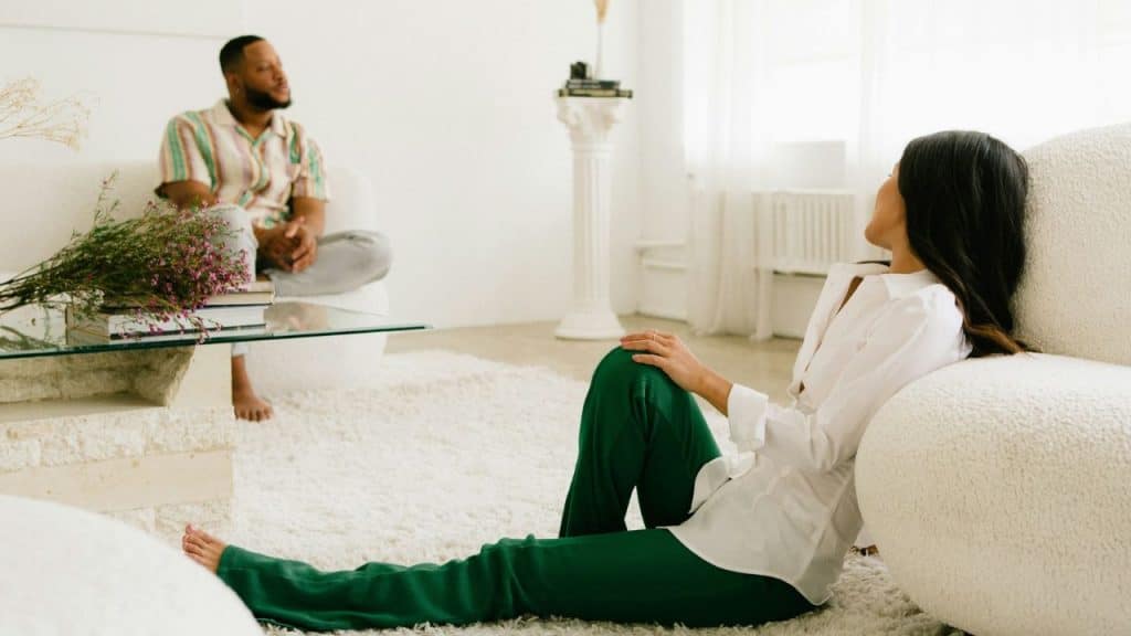 A man and a woman sit far apart in a white living room, looking away from each other.