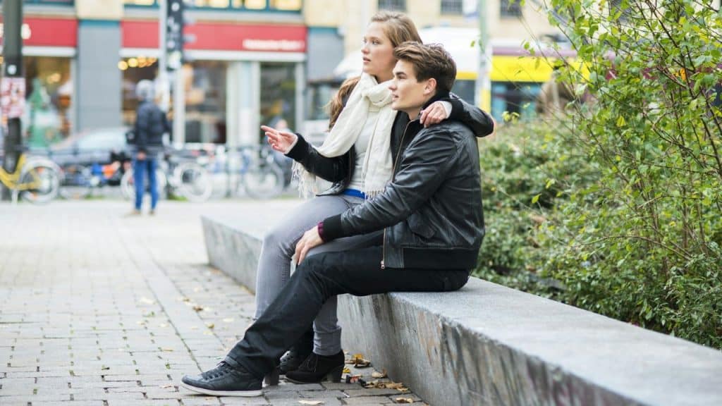 A young man and woman sit closely on a stone bench outdoors, looking away toward the street.