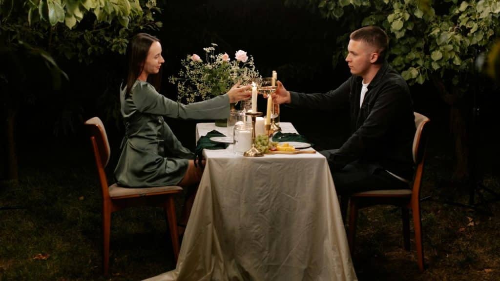 A man and woman toast with wine glasses at a candlelit table outdoors at night.