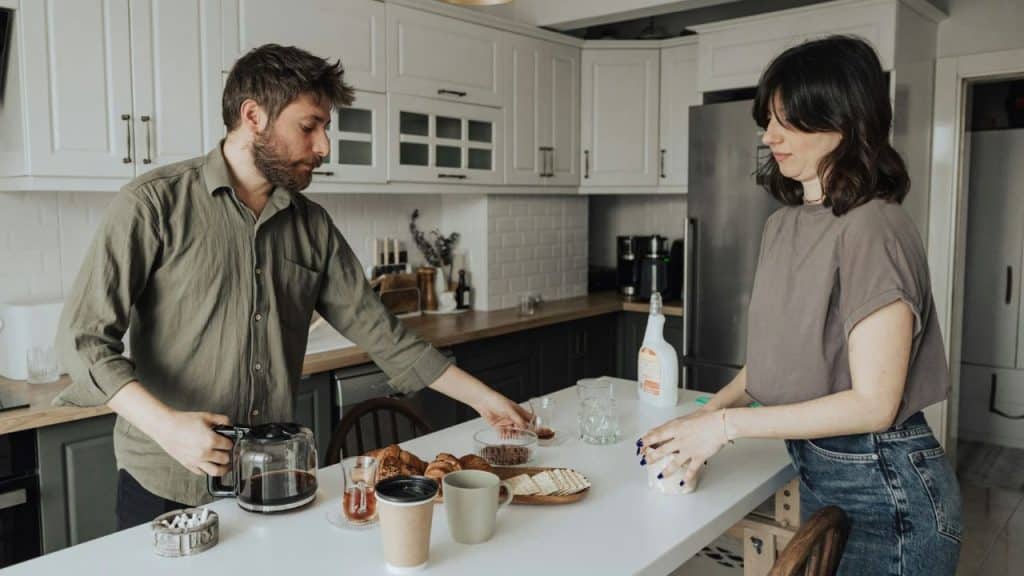 A man and a woman stand together preparing food and drinks on a white kitchen counter.