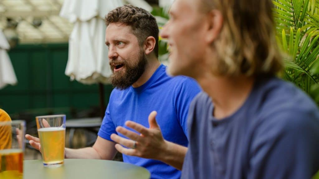 A bearded man in a blue shirt gestures while speaking to friends at an outdoor table with beer.