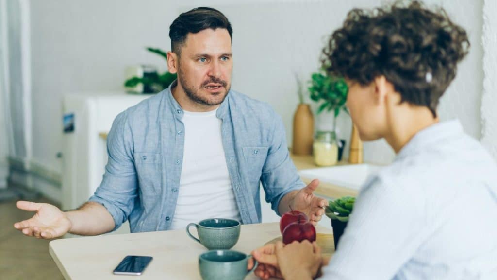 A man with a confused expression sits at a table, gesturing with both hands toward a woman.