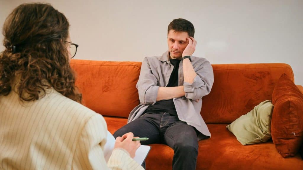 A cropped view of two people sitting on an orange couch against a white wall; one person is taking notes while the other sits with their hand on their forehead.