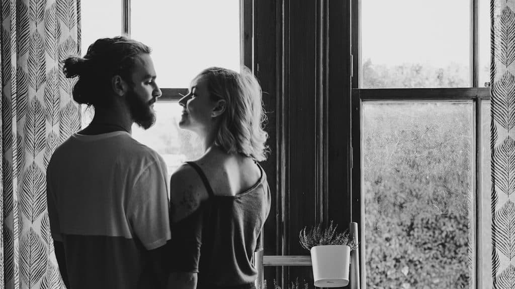 A bearded man and an attractive woman gazing lovingly into each other’s eyes and standing by a window.