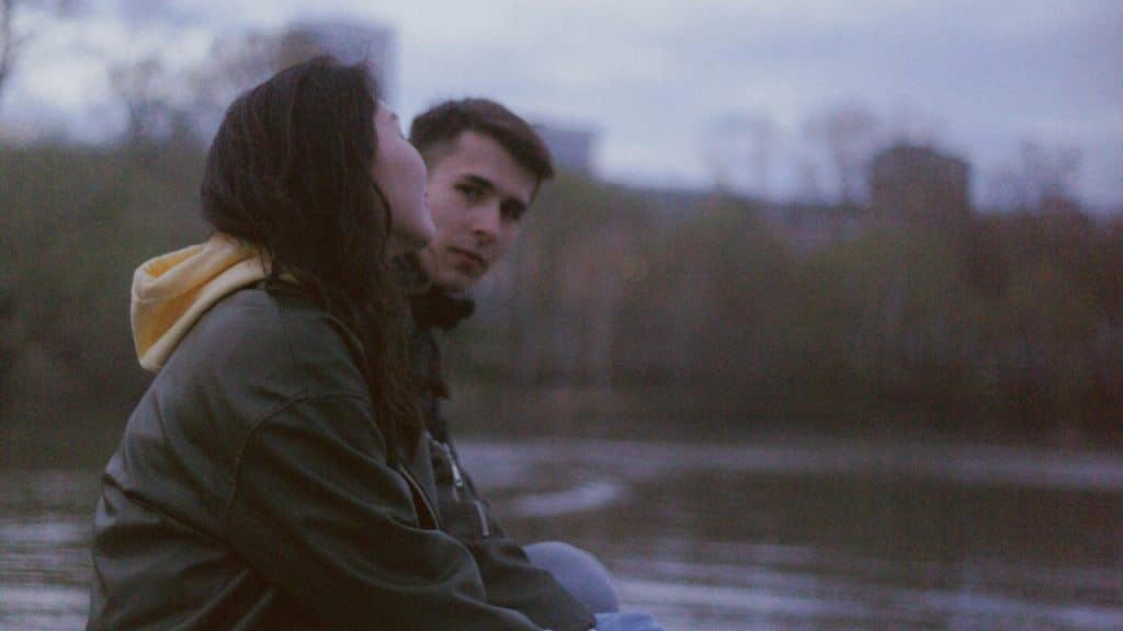 A man looking at his partner while sitting on a pier by a lake.