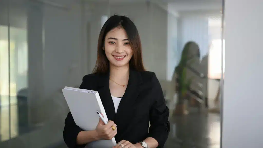 A smiling woman in business attire holding a file and pen.