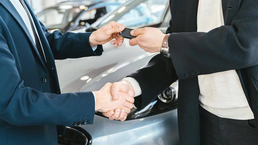 A person handing over car keys while shaking hands with another person at a dealership.