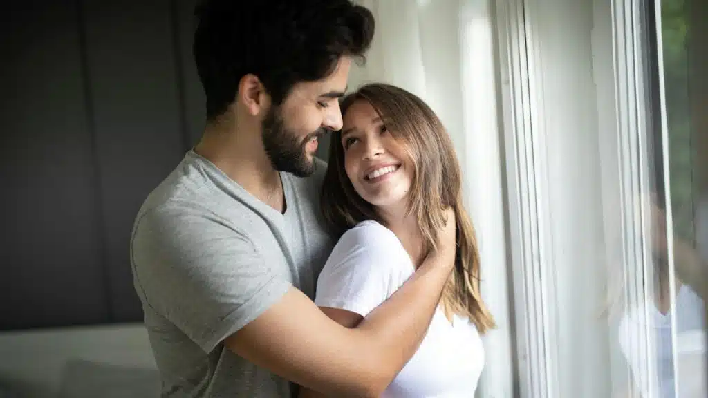 A couple smiling at each other and looking into each other’s eyes while the man holds the woman from behind.