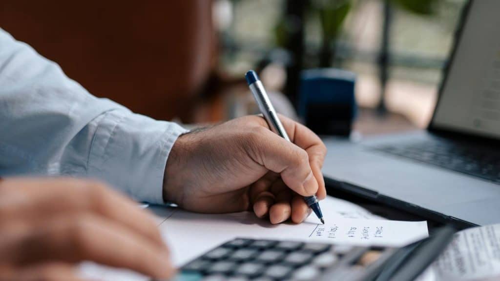 A person writing calculations on paper beside a laptop and calculator.
