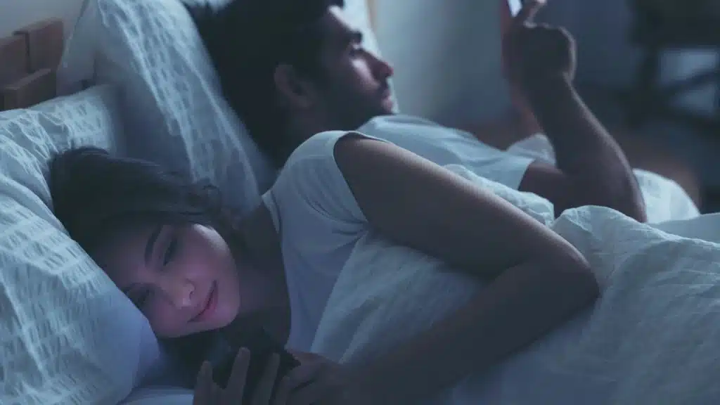 A man and woman lie in bed at night, facing away from each other, both looking at their phones.
