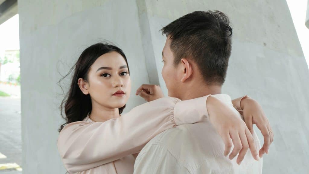 A young woman hugging a man from the front under a concrete structure, looking at the camera over his shoulder.